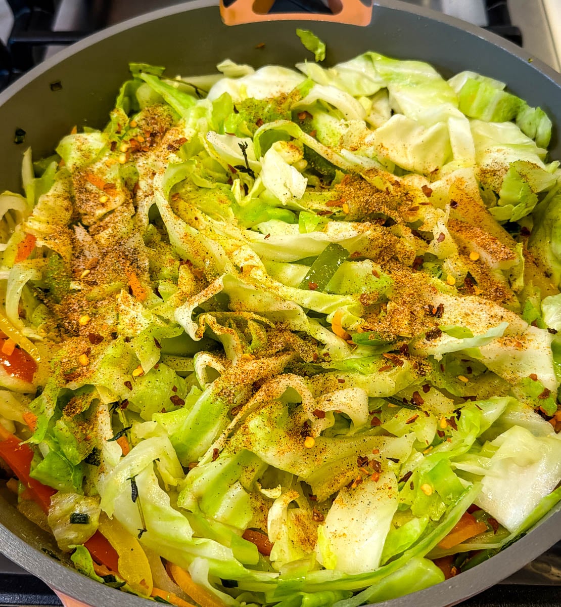 Chopped cabbage in a skillet topped with seasoning and red pepper flakes, ready to be cooked into Jamaican steamed cabbage.