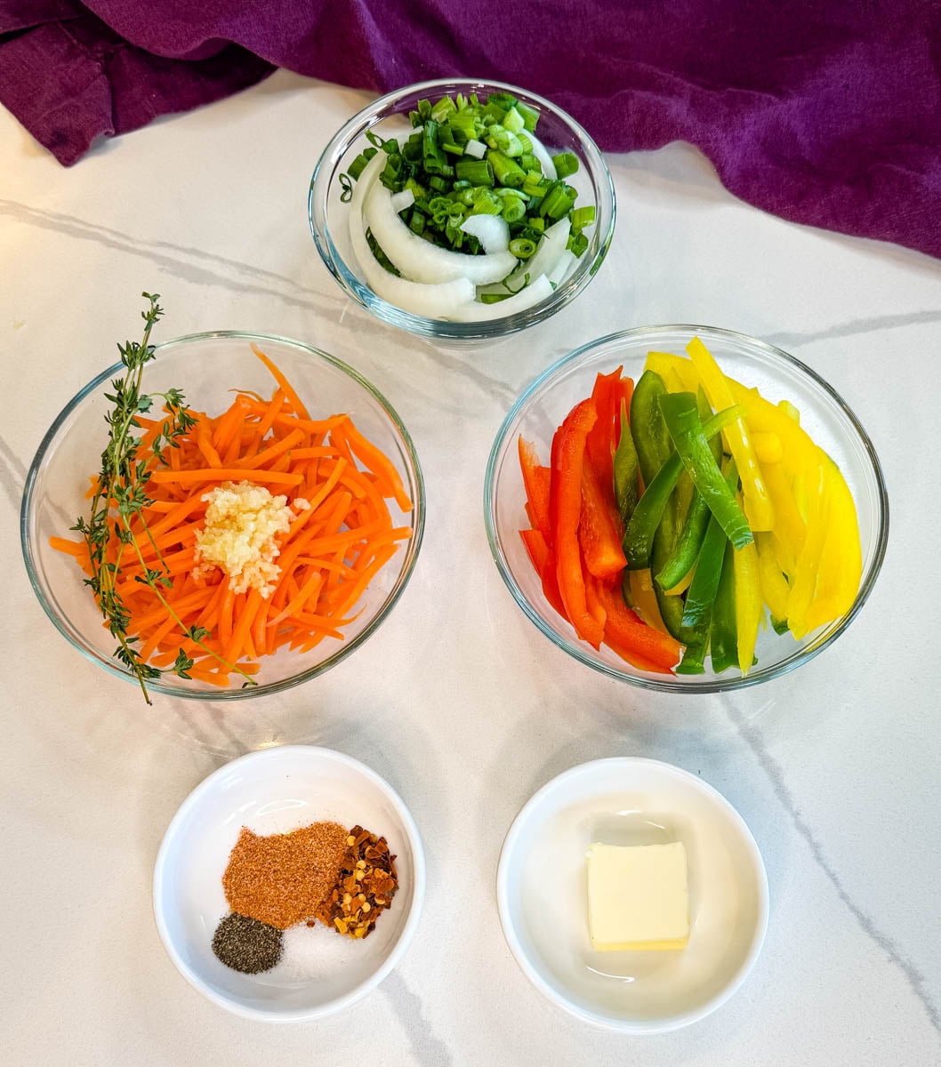 Ingredients for Jamaican steamed cabbage including sliced bell peppers, carrots, onions, green onions, garlic, thyme, butter, and seasoning in small bowls.