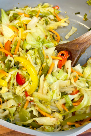 Jamaican steamed cabbage with bell peppers, carrots, onions, and herbs being stirred in a skillet with a wooden spoon.