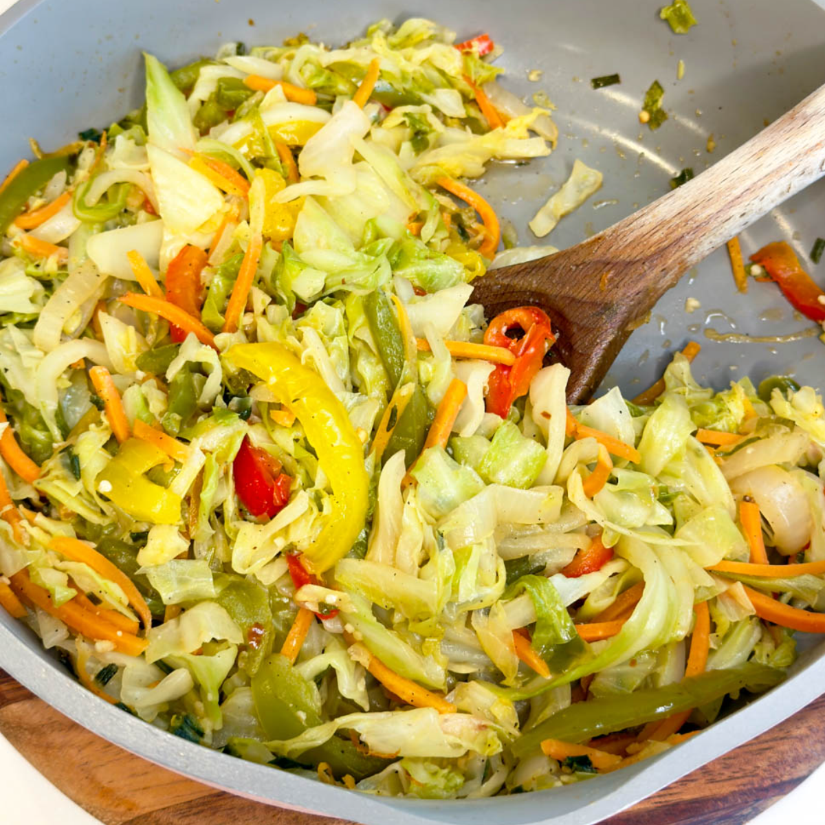 Jamaican steamed cabbage with bell peppers, carrots, onions, and herbs being stirred in a skillet with a wooden spoon.