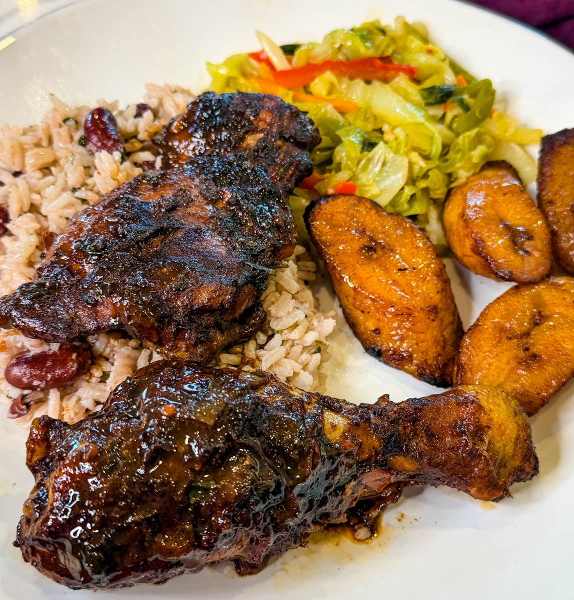 Plate of Jamaican brown stew chicken served with rice and peas, fried ripe plantains, and steamed cabbage.