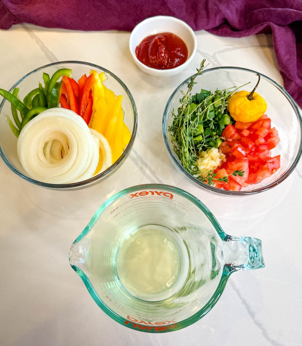 Bowls of sliced bell peppers, onions, green onions, garlic, thyme, diced tomatoes, and a whole scotch bonnet pepper with oil and ketchup for Jamaican brown stew chicken.