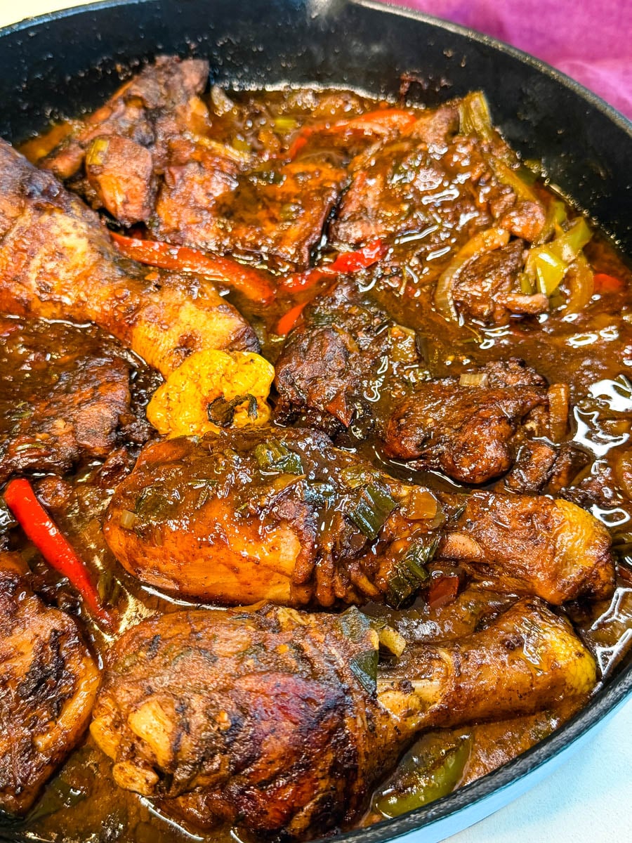 Close-up of brown stew chicken simmering in a rich, glossy gravy with peppers, onions, and a whole scotch bonnet pepper.