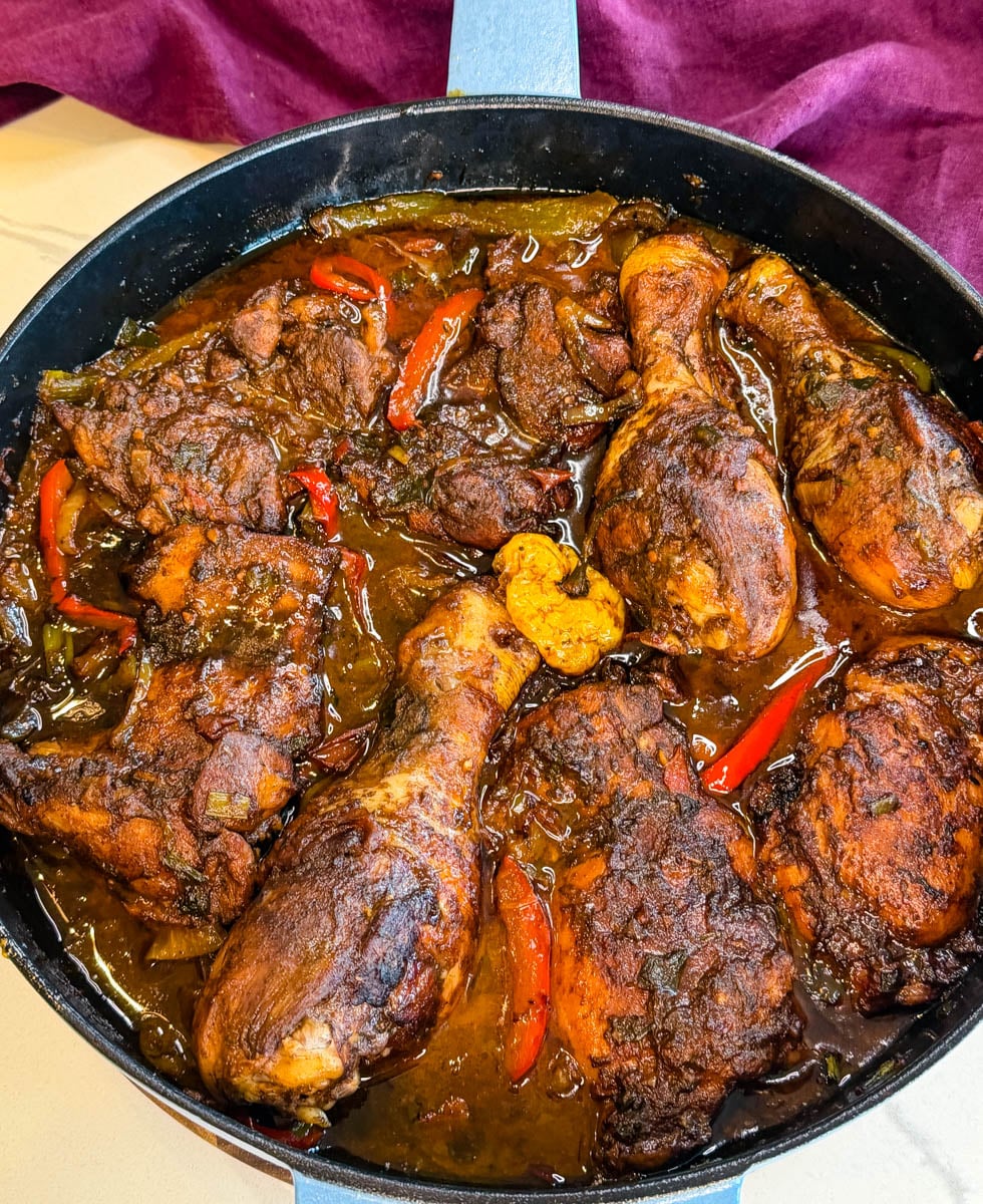 Overhead view of brown stew chicken in a skillet with thick brown gravy, bell peppers, onions, and herbs.
