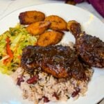 Jamaican brown stew chicken plated with rice and peas, caramelized plantains, and sautéed cabbage on the side.