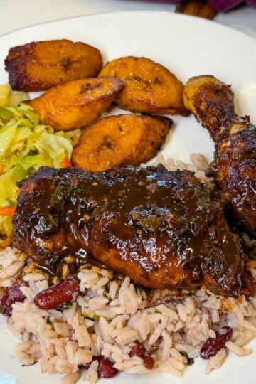 Jamaican brown stew chicken plated with rice and peas, caramelized plantains, and sautéed cabbage on the side.