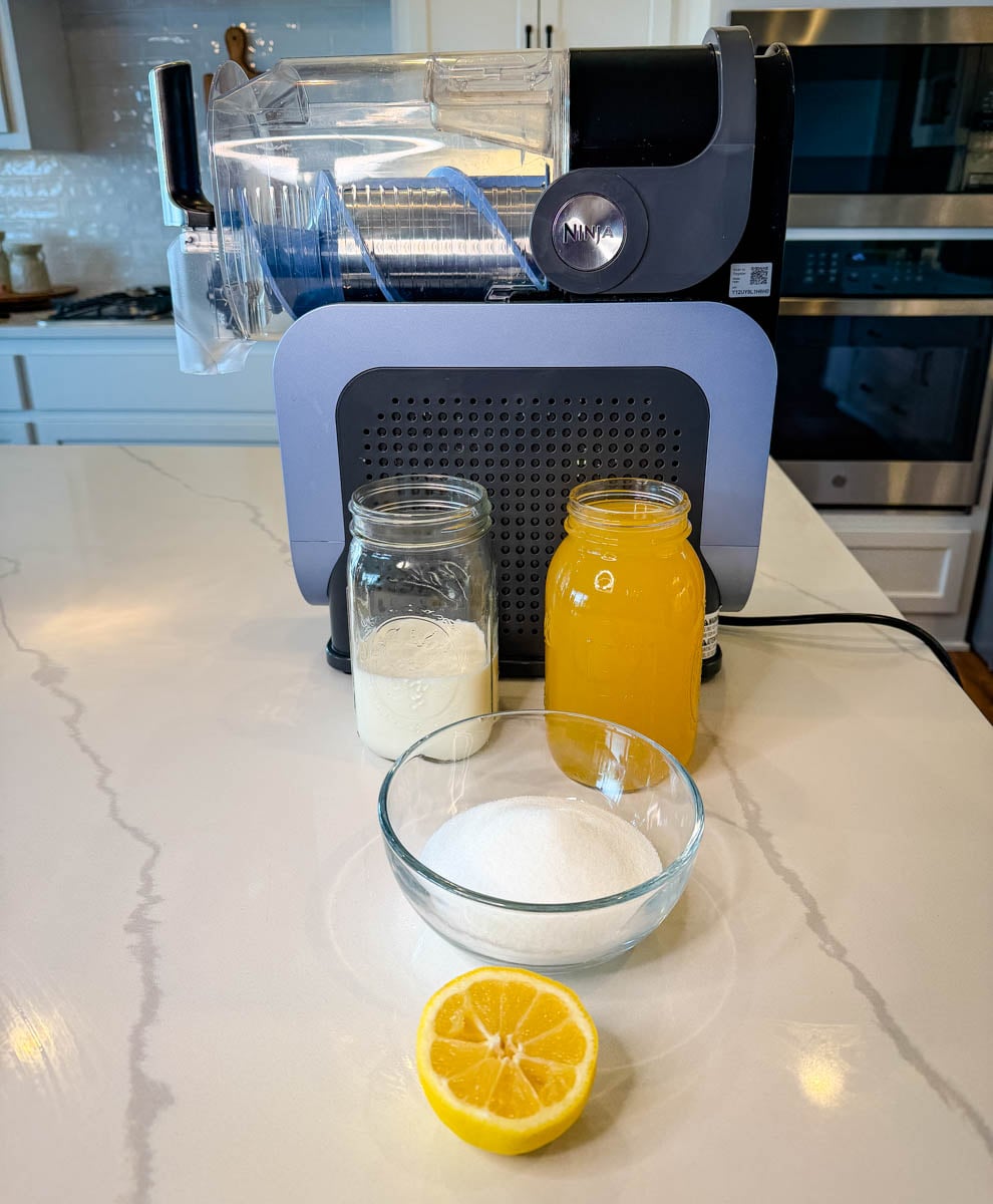Ingredients for Ninja Slushi Dole Whip including pineapple juice, milk, sugar, lemon, and Ninja Slushi machine on a kitchen counter