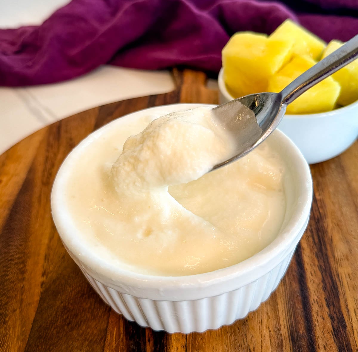 Spoon scooping creamy Ninja Slushi Dole Whip from a ramekin with fresh pineapple chunks in the background