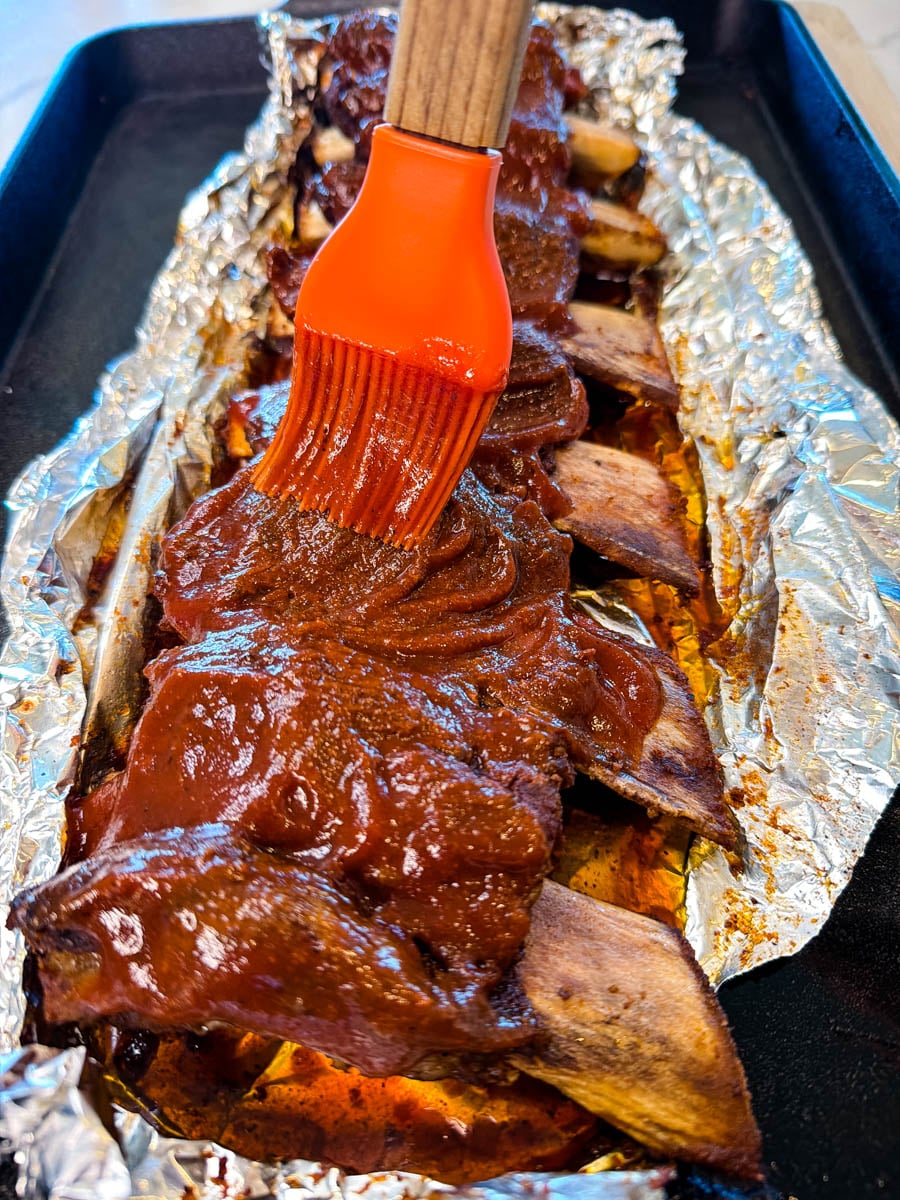 Brushing BBQ sauce over baked beef ribs in foil using a silicone brush.