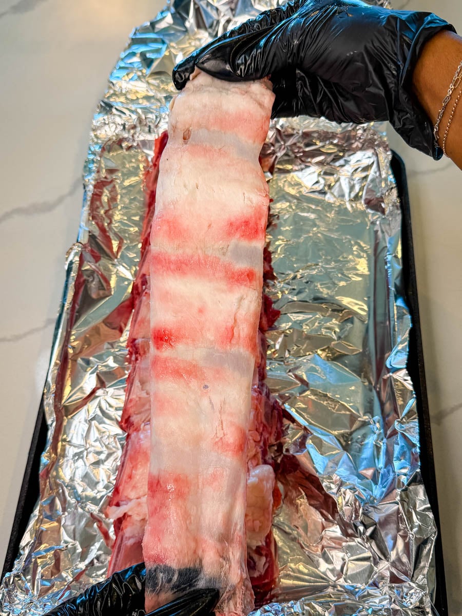 Removing the membrane from raw beef back ribs on a foil-lined baking sheet using gloved hands
