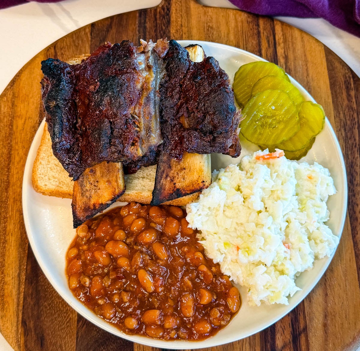 Close-up of baked beef ribs with BBQ sauce served with baked beans, coleslaw, pickles, and white bread.