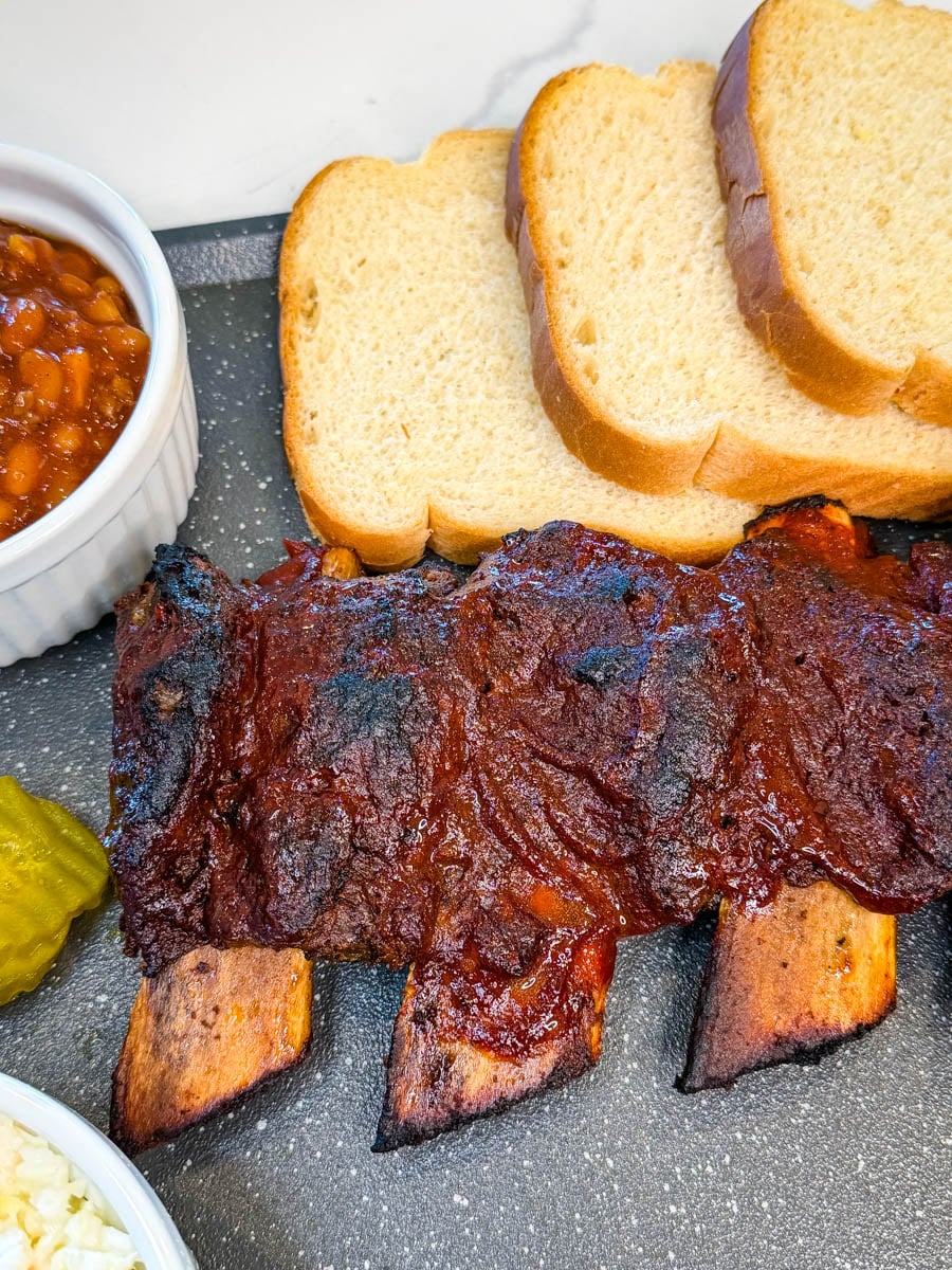 Full rack of oven baked beef ribs with a rich BBQ sauce glaze on a serving tray with bread and sides.