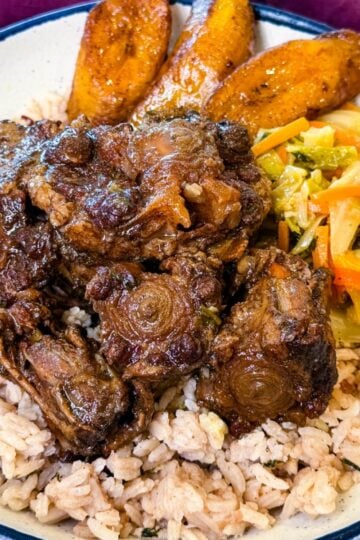 Jamaican oxtail plated over rice and peas with golden fried plantains and colorful steamed vegetables.