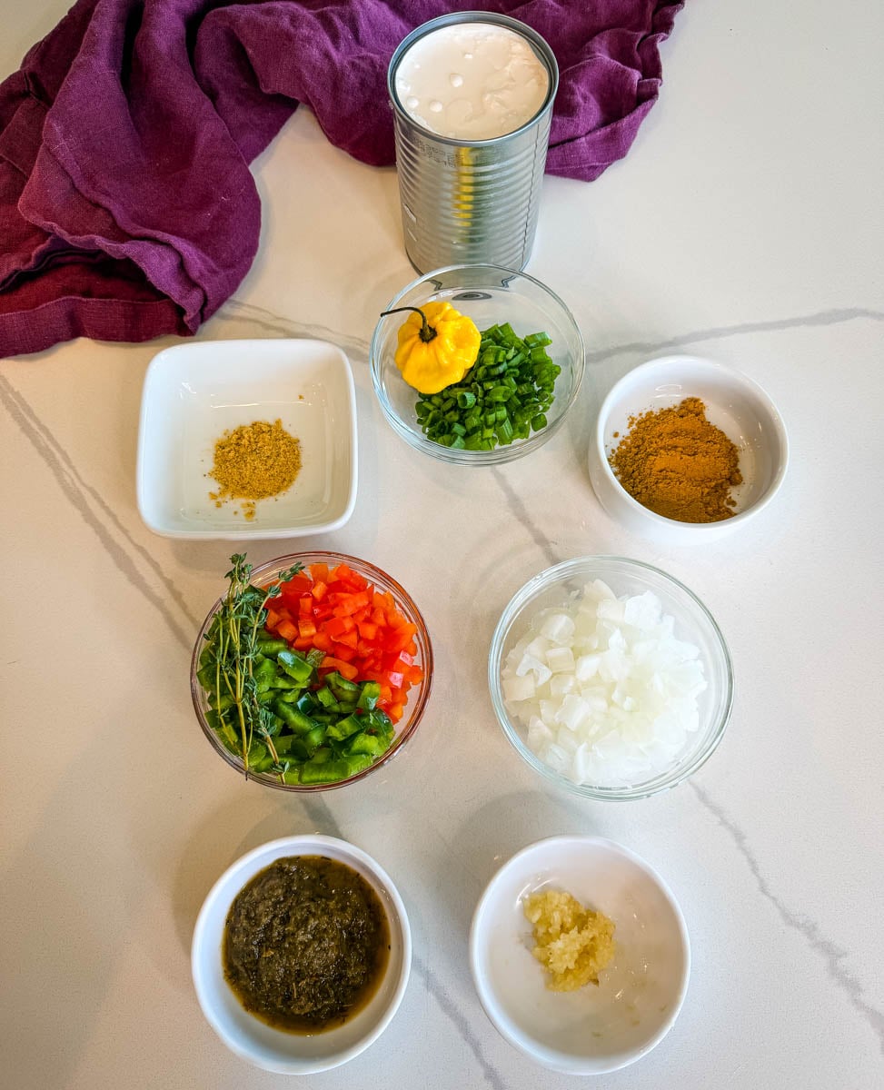 Ingredients for Jamaican curry shrimp arranged in small bowls including coconut milk, curry powder, green seasoning, Scotch bonnet pepper, onions, garlic, bell peppers, thyme, and green onions.