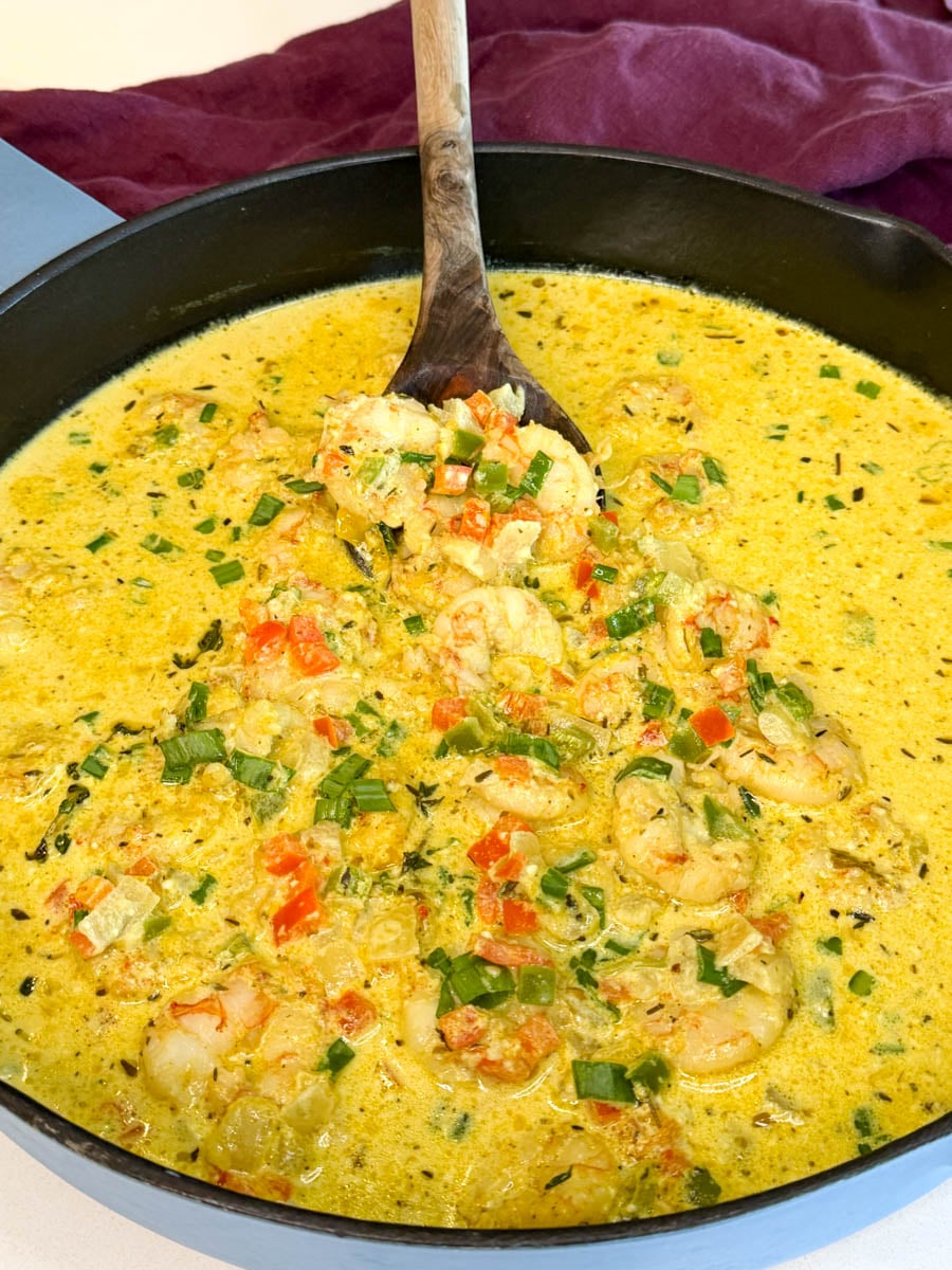 Jamaican curry shrimp simmering in a creamy coconut curry sauce with bell peppers, onions, thyme, and green onions in a black skillet with a wooden spoon.