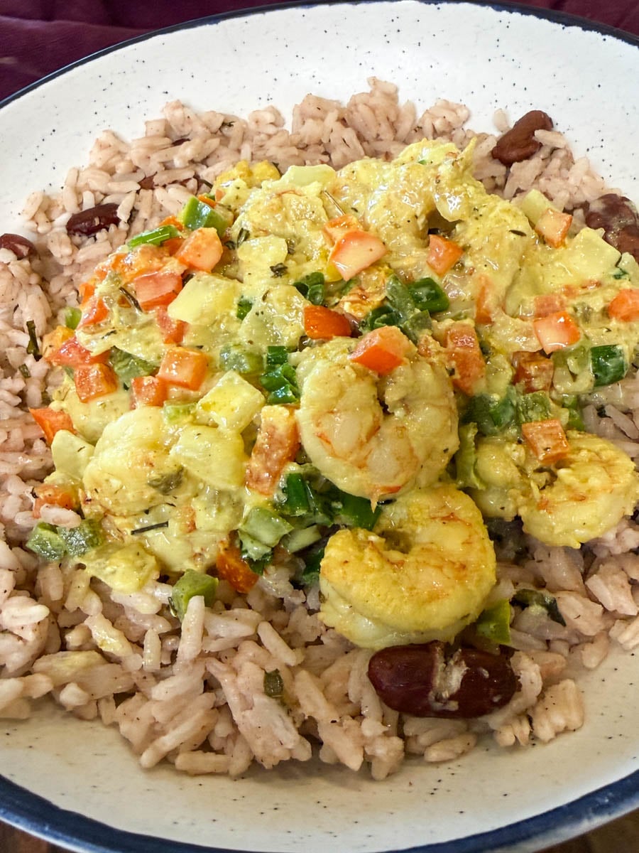 Jamaican curry shrimp served over rice and peas in a white bowl, topped with a creamy coconut curry sauce, bell peppers, onions, green onions, and tender shrimp.