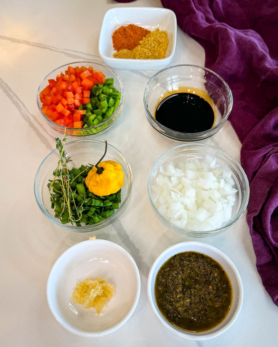 Small bowls filled with chopped bell peppers, onions, green seasoning, garlic, fresh thyme, Scotch bonnet pepper, browning sauce, and Jamaican seasonings arranged on a countertop for making Jamaican oxtail.