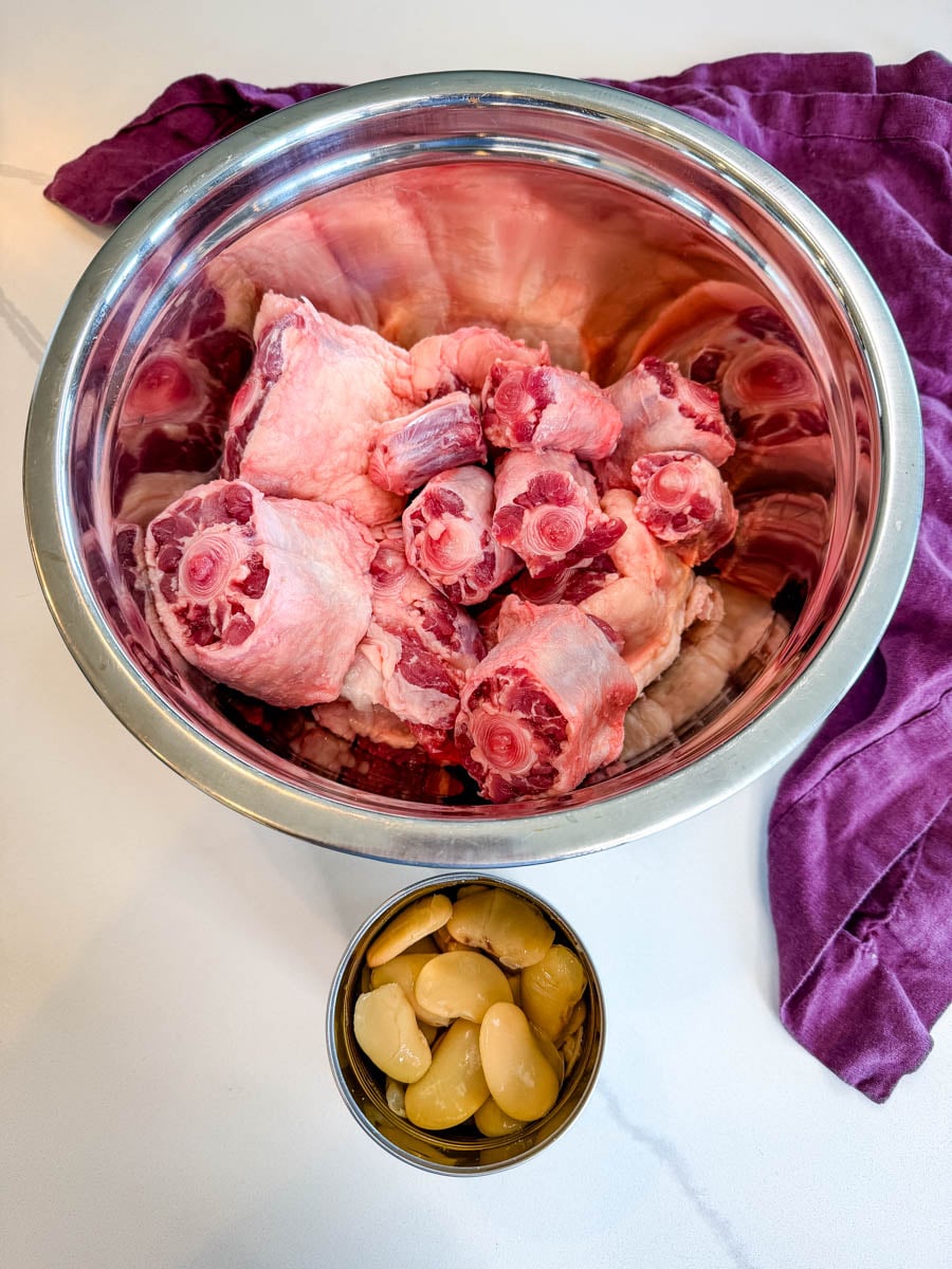 Raw oxtail pieces in a large metal bowl alongside a can of butter beans, ready to be seasoned and cooked for Jamaican oxtail.