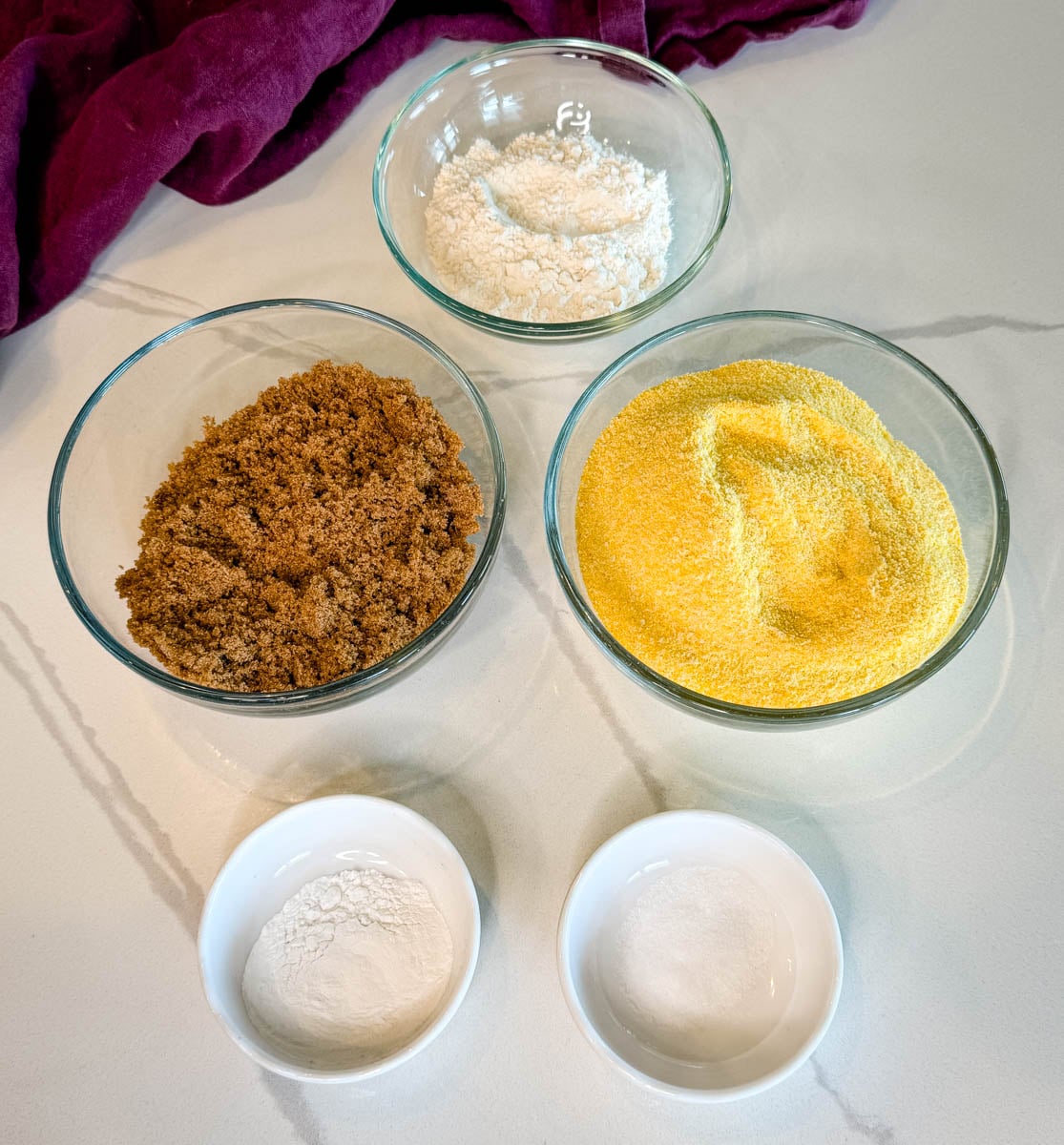 Bowls of cornmeal, brown sugar, flour, baking powder, and salt arranged on a countertop for making sweet honey butter cornbread.