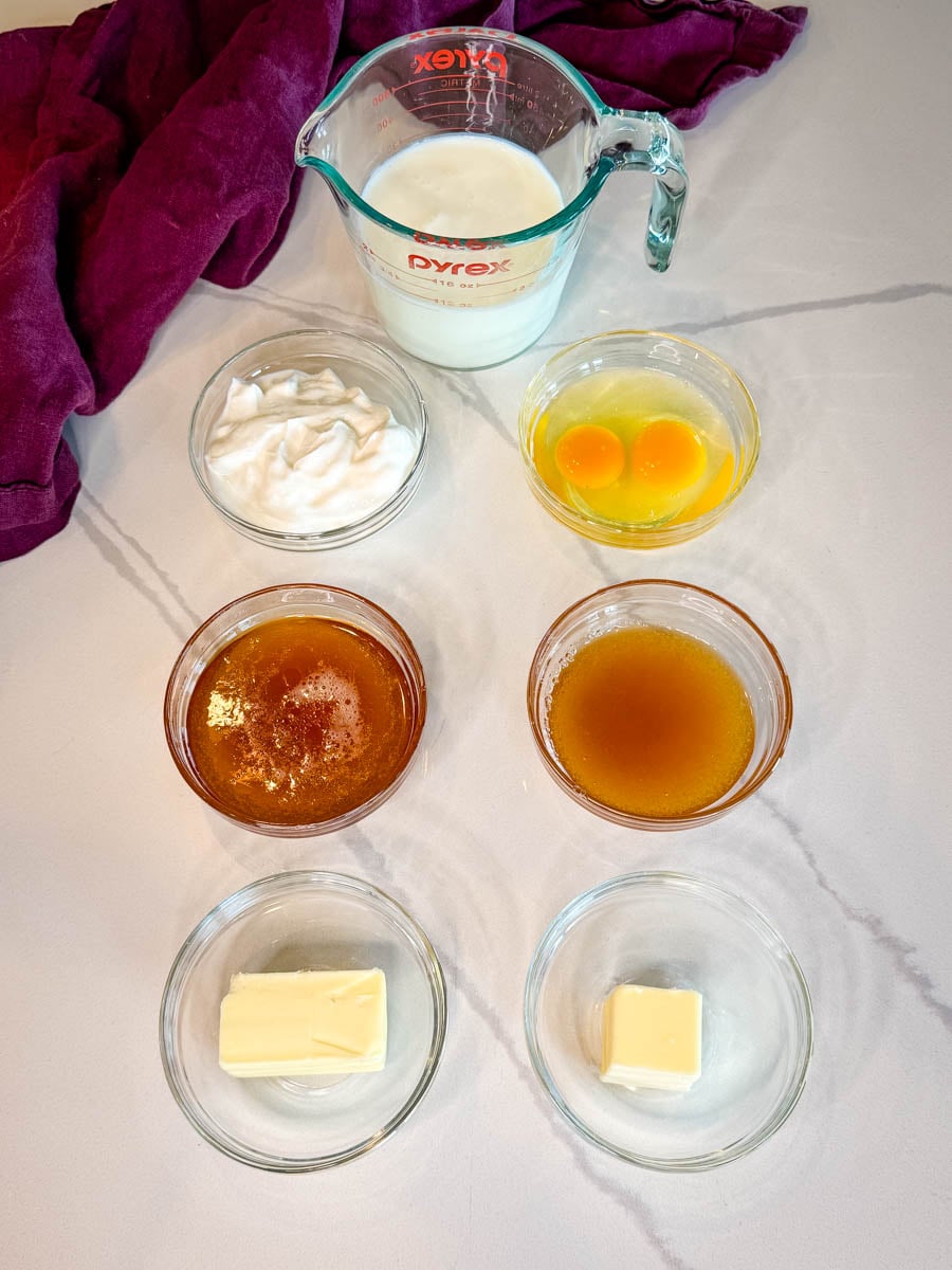 Ingredients for sweet cornbread including milk, sour cream, eggs, honey, vanilla extract, and butter measured out in glass bowls.