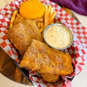 Crispy fried cod served with fries, tartar sauce, and hot water cornbread in a red checkered basket.