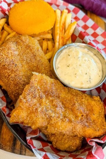 Crispy fried cod served with fries, tartar sauce, and hot water cornbread in a red checkered basket.