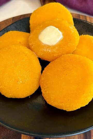 Golden hot water cornbread patties stacked on a black plate with melted butter on top and a purple cloth in the background.