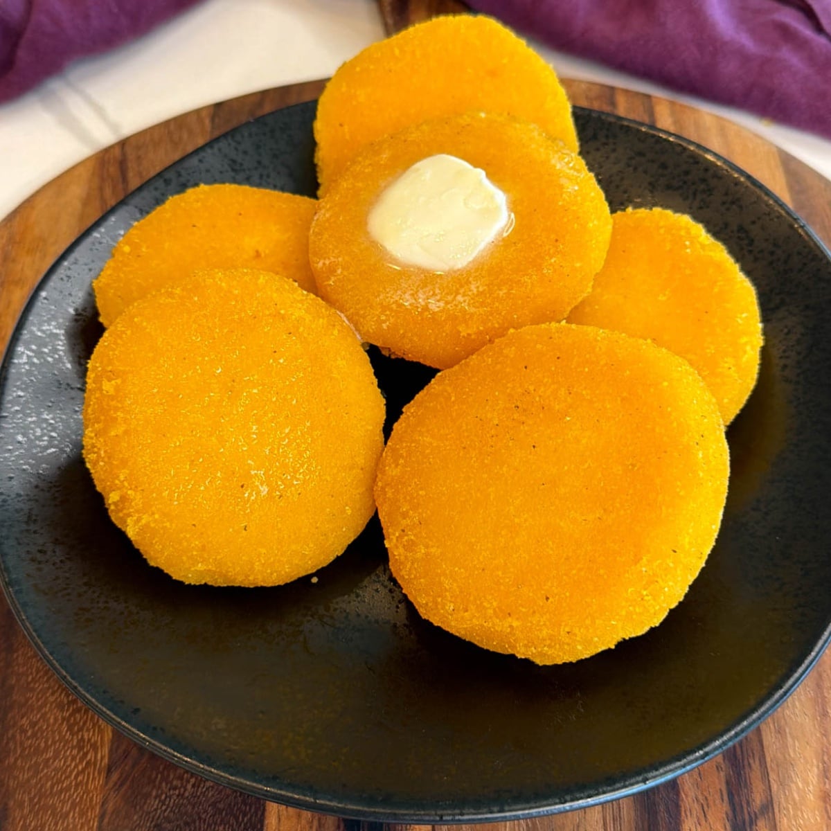 Golden hot water cornbread patties stacked on a black plate with melted butter on top and a purple cloth in the background.