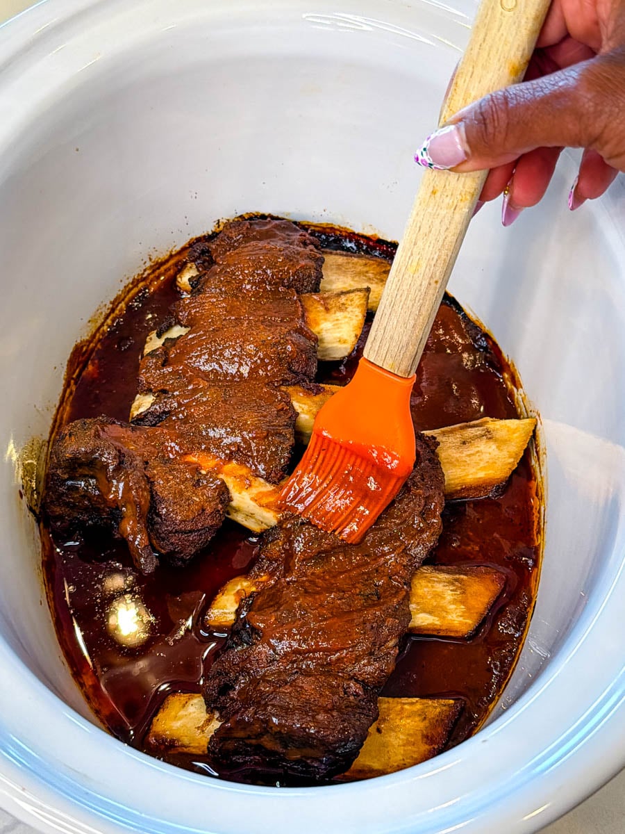 Beef ribs in the Crockpot being brushed with BBQ sauce using a silicone basting brush.