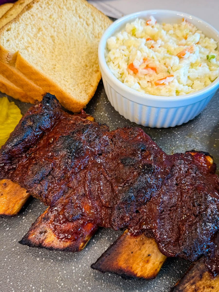BBQ beef ribs served with coleslaw, pickles, and white bread on a plate.
