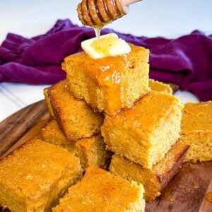 Close-up of sweet honey butter cornbread stacked high with butter melting on top while honey is drizzled from a honey dipper.