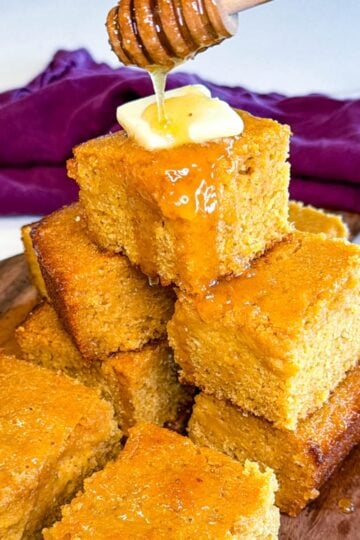 Close-up of sweet honey butter cornbread stacked high with butter melting on top while honey is drizzled from a honey dipper.