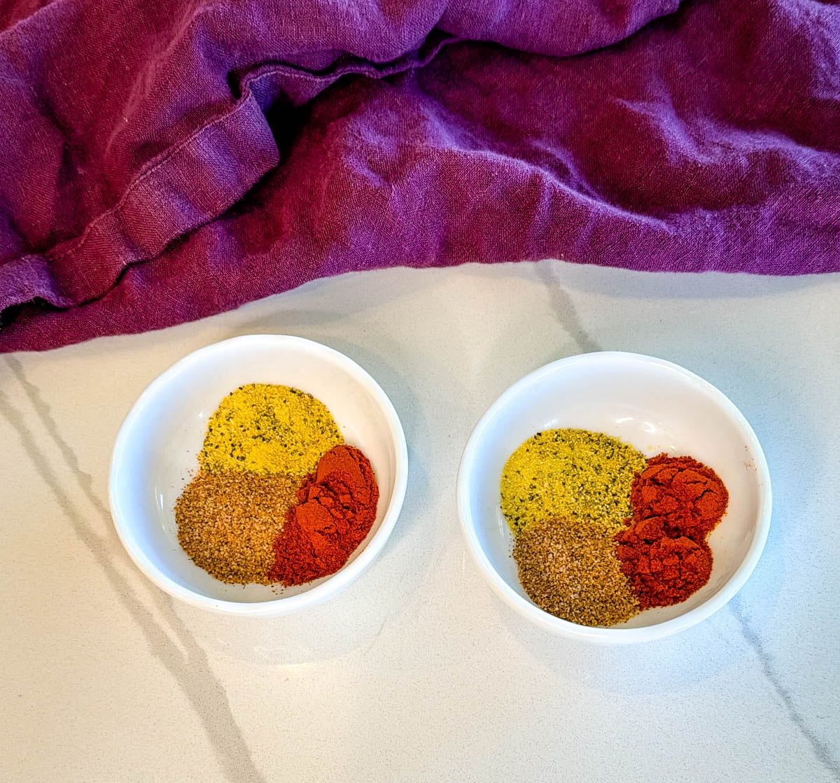 Small bowls filled with seasoning blends for fried cod including paprika, lemon pepper seasoning, and spices on a countertop with a purple cloth backdrop.