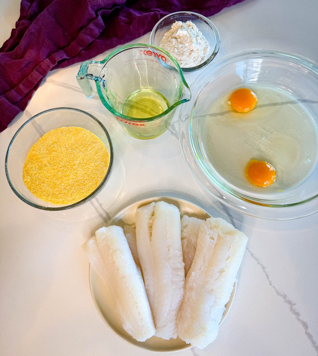 Ingredients for fried cod including raw cod fillets, eggs, flour, cornmeal, oil, and seasonings arranged on a countertop.