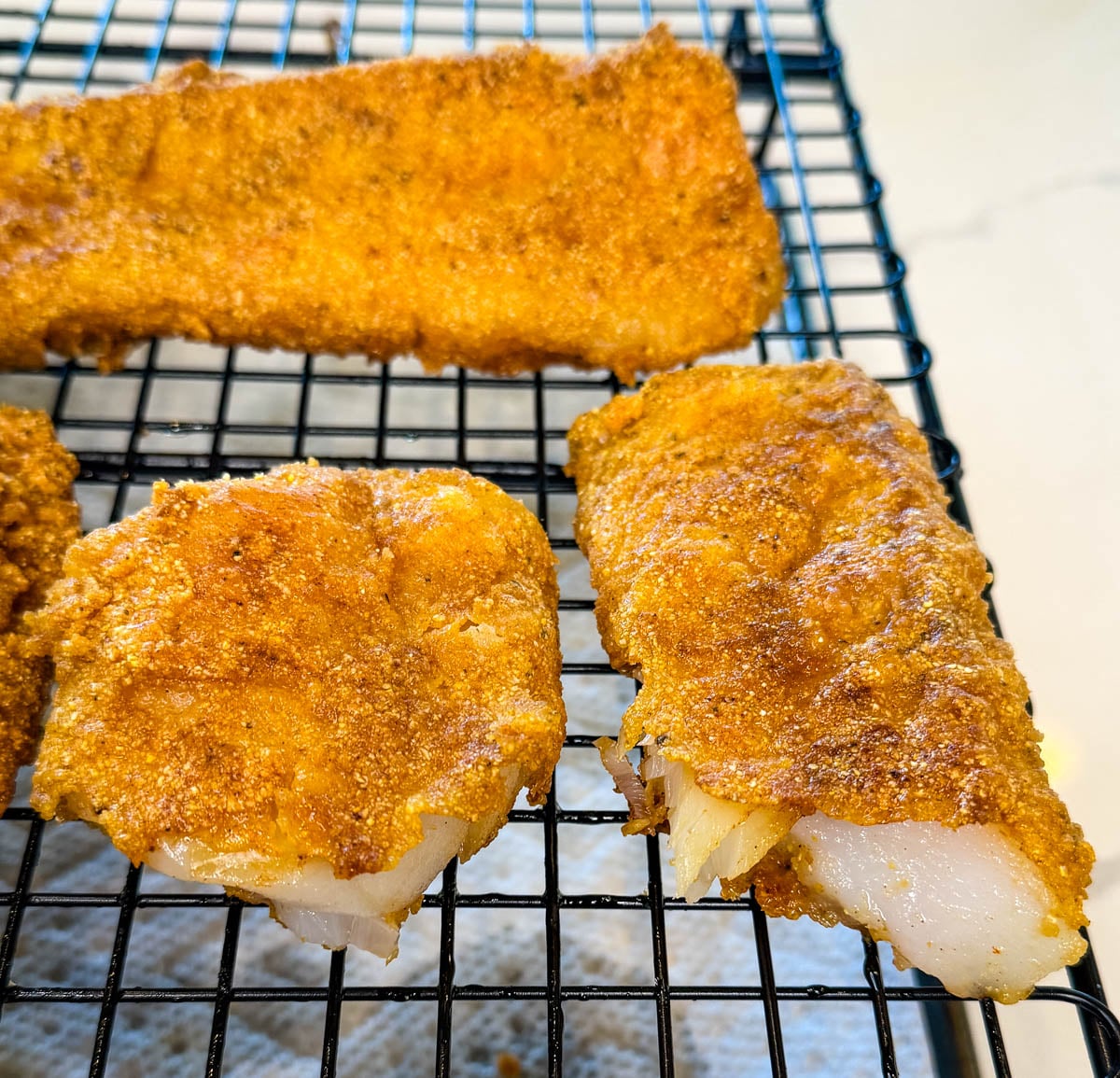 Freshly fried cod fillets cooling on a wire rack with golden crispy breading and flaky white fish inside.