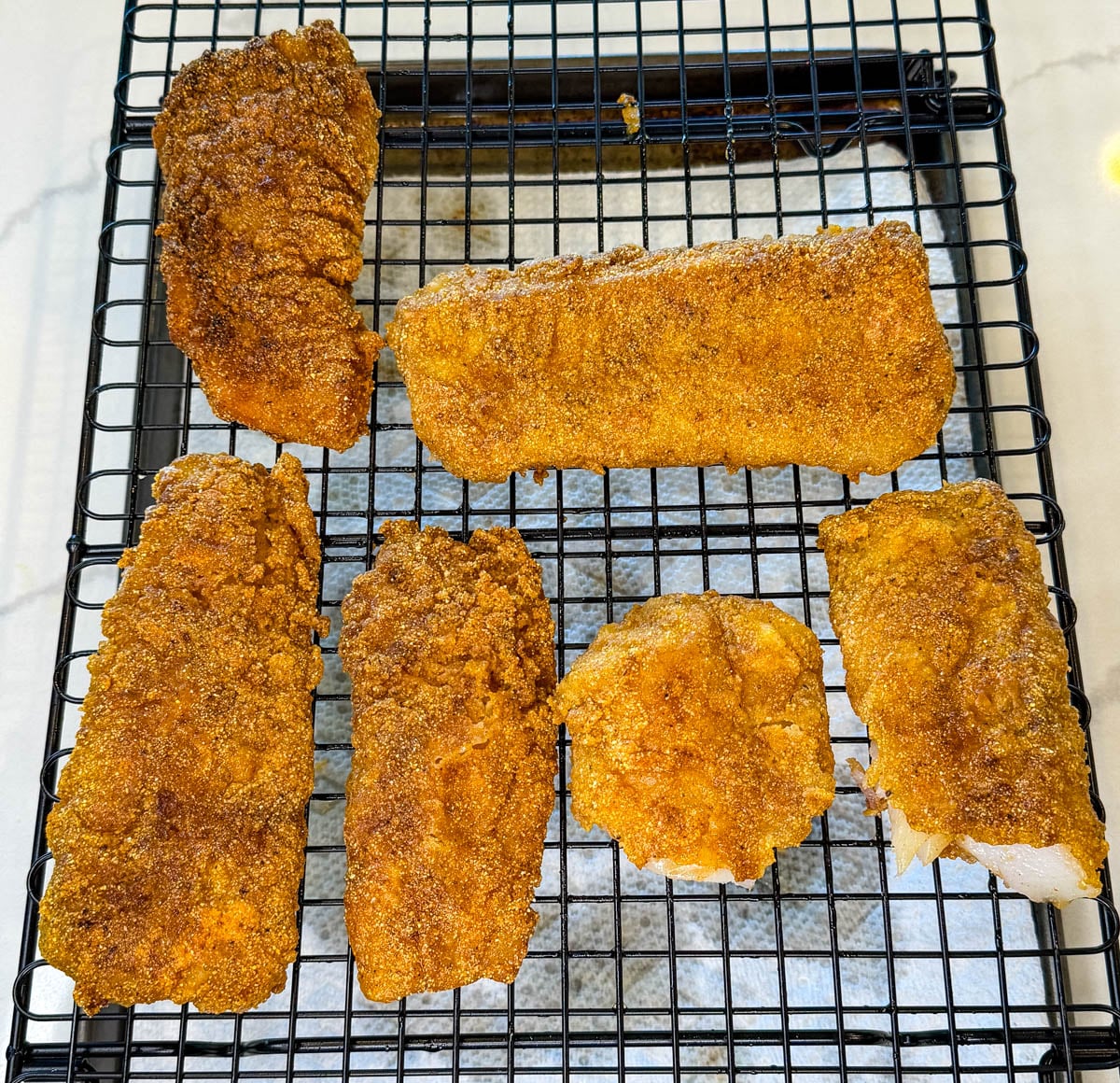 Crispy fried cod fillets cooling on a wire rack after frying, showing golden cornmeal breading and flaky white fish inside.