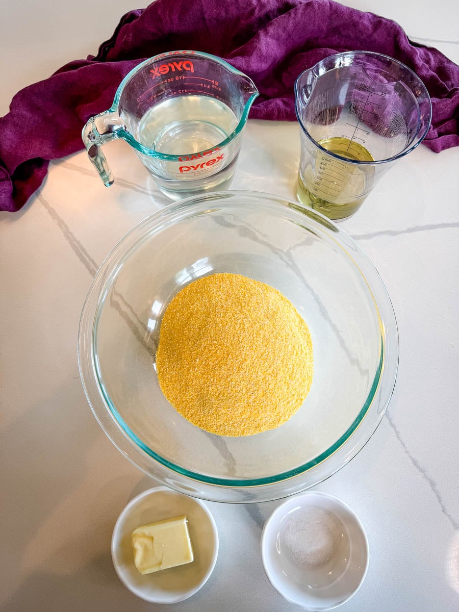 Ingredients for hot water cornbread including cornmeal, hot water, oil, butter, and salt arranged on a countertop beside a purple cloth.