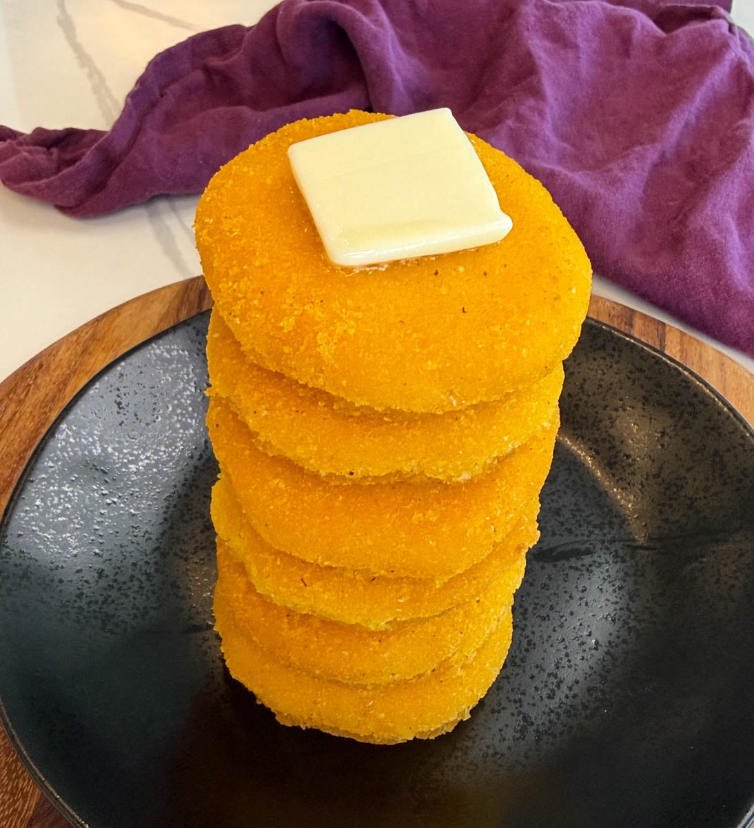 Tall stack of crispy hot water cornbread on a black plate topped with a square of butter.