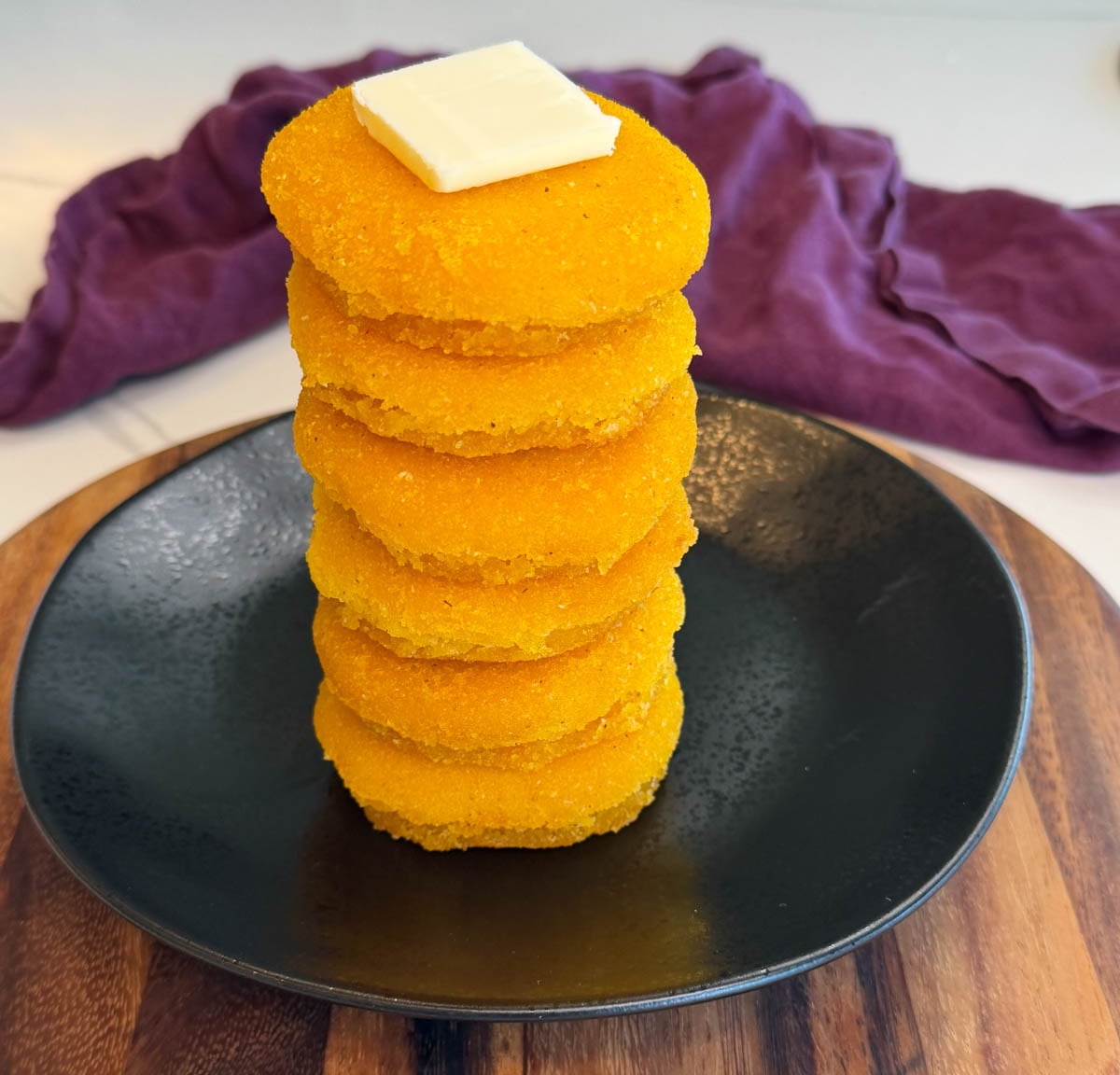 Vertical stack of Southern hot water cornbread served on a black plate with butter and a purple cloth backdrop.
