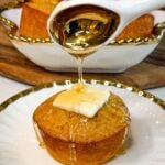 Freshly baked honey butter cornbread muffin topped with butter while honey is drizzled over the top, with additional muffins in the background.