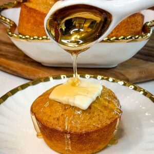 Freshly baked honey butter cornbread muffin topped with butter while honey is drizzled over the top, with additional muffins in the background.