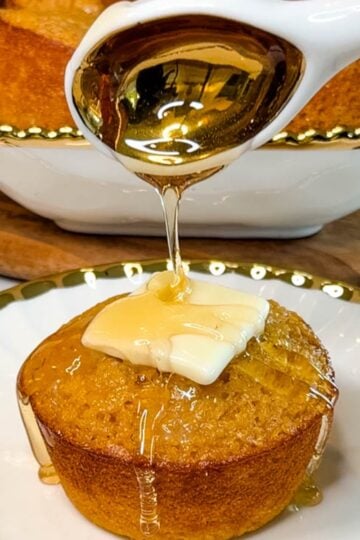 Freshly baked honey butter cornbread muffin topped with butter while honey is drizzled over the top, with additional muffins in the background.