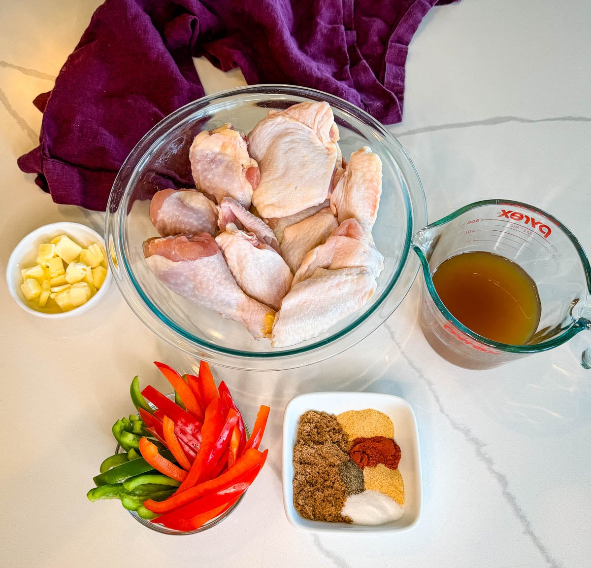 Raw chicken thighs, wings, and drumsticks with sliced bell peppers, butter, broth, and seasonings arranged on a countertop for baked chicken.