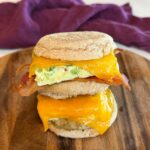 Stack of meal prep breakfast sandwiches made with English muffins, eggs, melted cheddar cheese, and bacon or sausage on a wooden serving board.