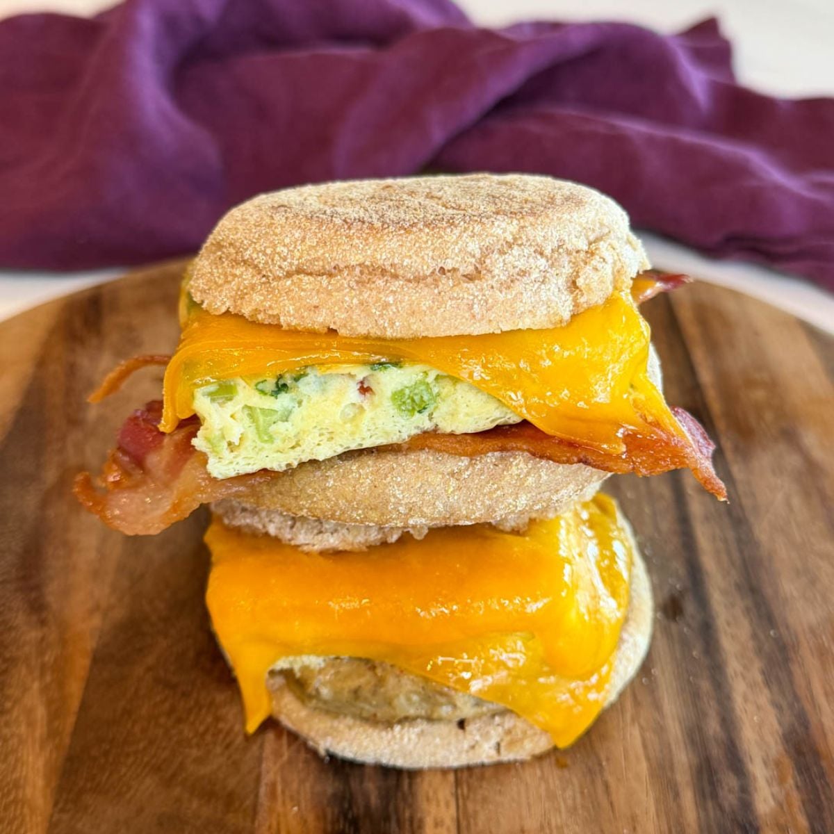 Stack of meal prep breakfast sandwiches made with English muffins, eggs, melted cheddar cheese, and bacon or sausage on a wooden serving board.