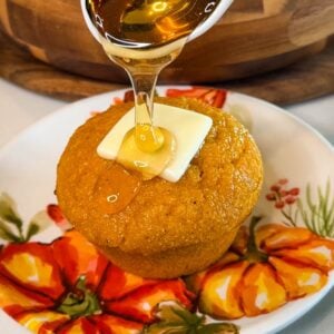 Honey being drizzled over a warm pumpkin cornbread muffin topped with a pat of butter on a fall-themed pumpkin plate.