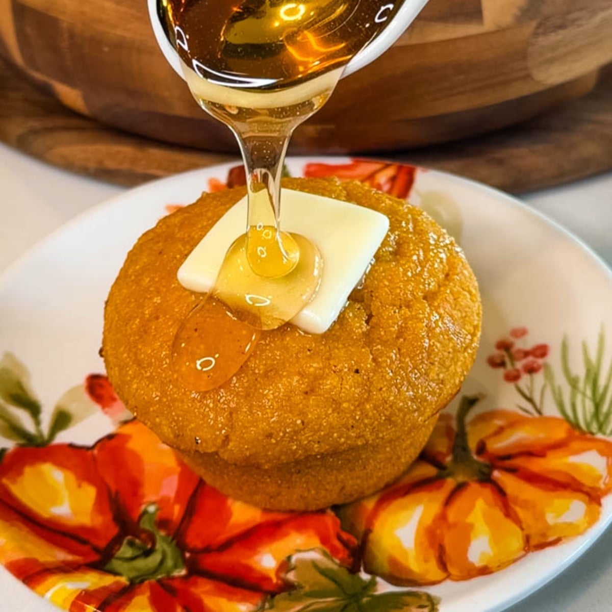 Honey being drizzled over a warm pumpkin cornbread muffin topped with a pat of butter on a fall-themed pumpkin plate.