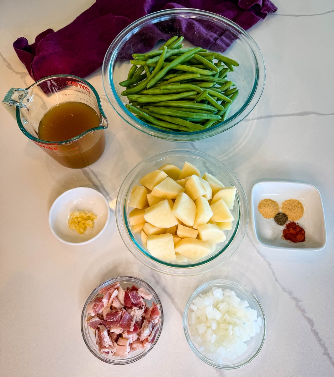 Ingredients for Southern green beans and potatoes arranged on a countertop, including fresh green beans, russet potatoes, bacon, onions, garlic, broth, and seasonings.