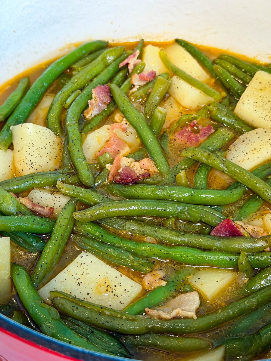 Southern-style green beans and potatoes cooking in a rich broth with pieces of bacon and cracked black pepper.