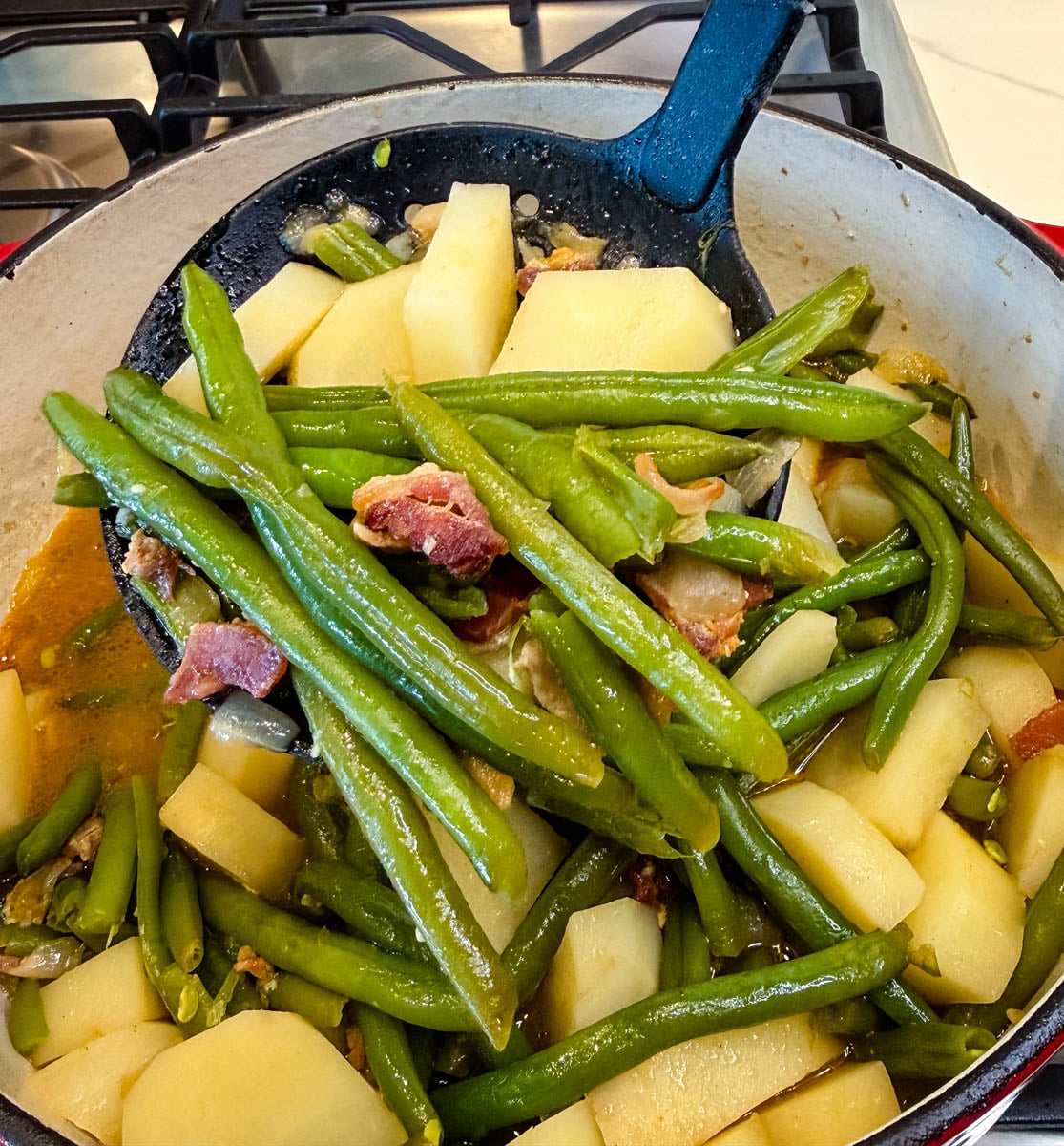 Spoonful of Southern green beans, potatoes, and bacon lifted from a Dutch oven filled with seasoned broth.
